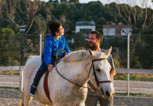 Criança a sorrir enquanto monta um cavalo branco, acompanhada por um terapeuta durante uma sessão de Terapia Assistida por Animais, num ambiente ao ar livre e descontraído.